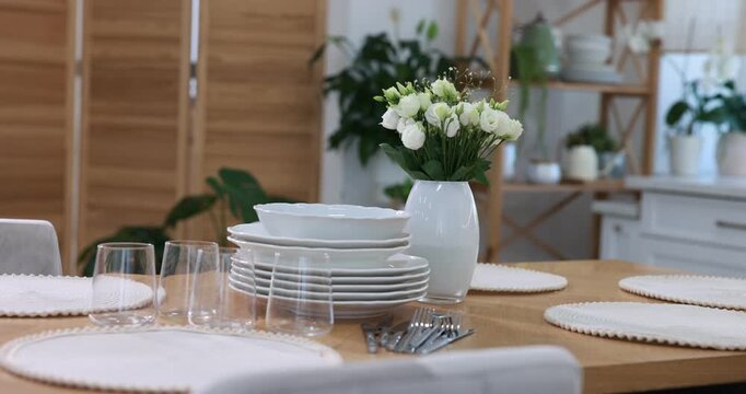 Stylish setting with vase of flowers, plates and glasses on wooden table in kitchen. Camera moving left