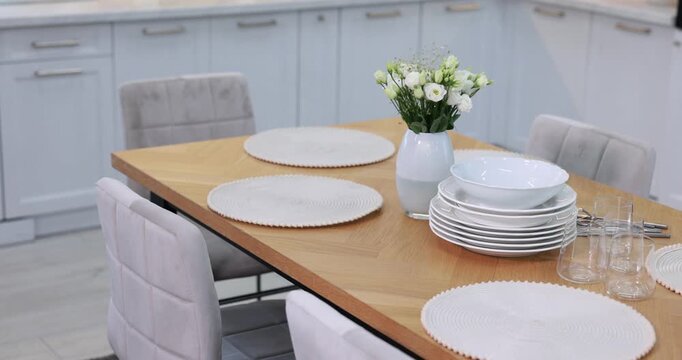 Stylish setting with vase of flowers, plates and glasses on wooden table in kitchen. Camera moving left