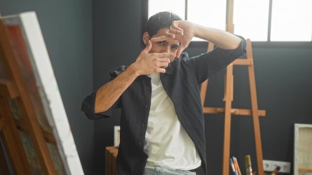 Young hispanic man frames a canvas with hands and fingers, gesturing composition beside an easel in an art studio; concentration.