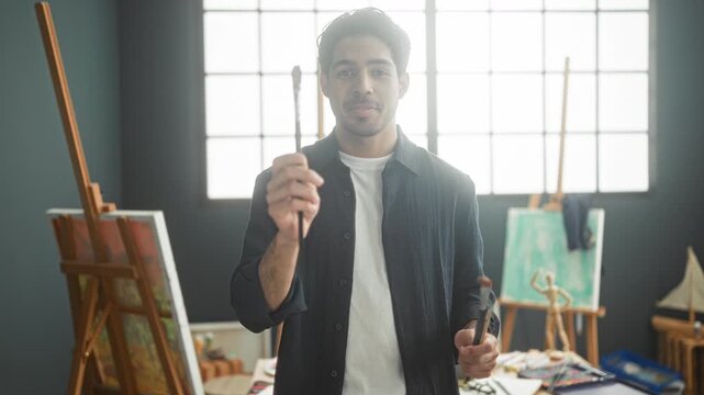 Man holding paintbrushes and gesturing with hands in a bright painting studio by an easel, canvas, palette and wooden mannequin; focused creativity.