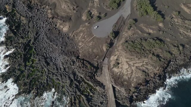 Capelinhos coastal area on Faial Island, Azores, Portugal, showing a causeway crossing a rugged rocky shoreline with Atlantic waves breaking on both sides, top-down aerial panning view
