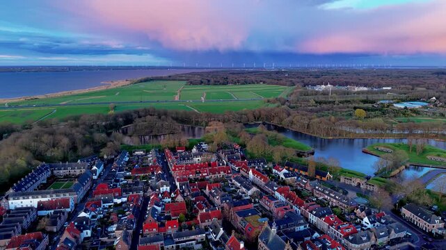 Beautiful high altitude aerial panoramic perspective of historic European town red brick roofs moat canal lake coast wind turbines and vibrant pink sunset purple clouds during twilight dusk
