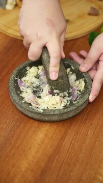 Close-up of hands grinding shallots and garlic using a traditional stone mortar and pestle on a wooden table.