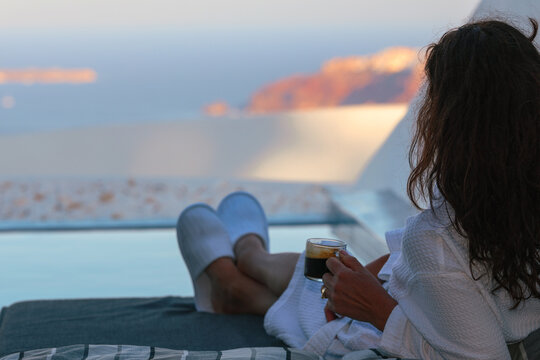 Woman relaxing beside a private infinity pool while enjoying a morning coffee overlooking the caldera in Santorini, Greece. She is wearing a robe while absorbing the view..