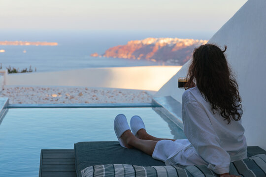 Woman relaxing beside a private infinity pool while enjoying a morning coffee overlooking the caldera in Santorini, Greece. She is wearing a robe while absorbing the view..