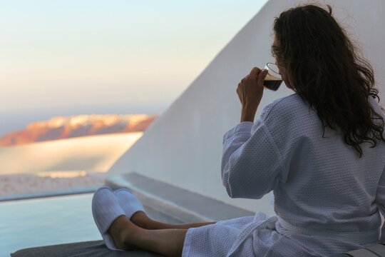 Woman relaxing beside a private infinity pool while enjoying a morning coffee overlooking the caldera in Santorini, Greece. She is wearing a robe while absorbing the view..
