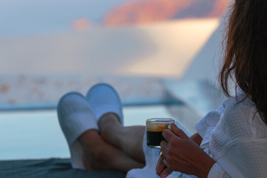 Woman relaxing beside a private infinity pool while enjoying a morning coffee overlooking the caldera in Santorini, Greece. She is wearing a robe while absorbing the view..