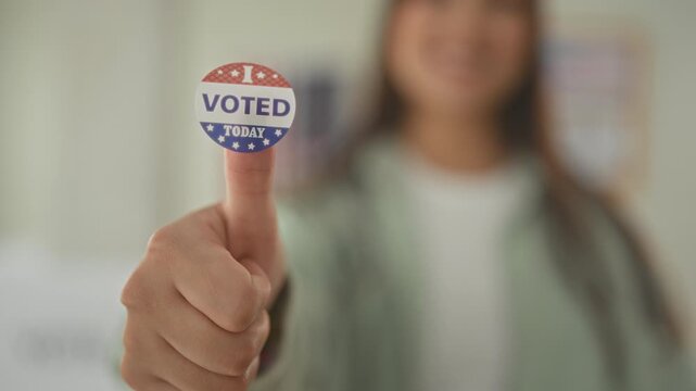 Young hispanic woman holds 'i voted today' sticker on raised thumb in a polling building, smiling and offering the sticker toward camera; civic pride.