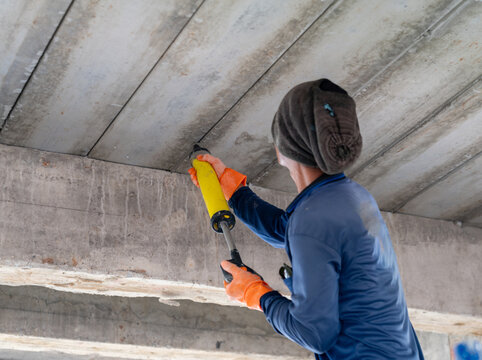 Construction worker applying sealant or polyurethane foam into joint gaps of concrete ceiling slabs using a manual applicator gun.