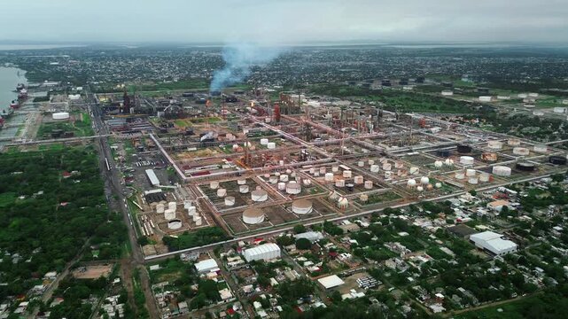 Aerial drone footage of petroleum refinery with smoke emissions from industrial stacks in green zone with Tampico cityscape background
