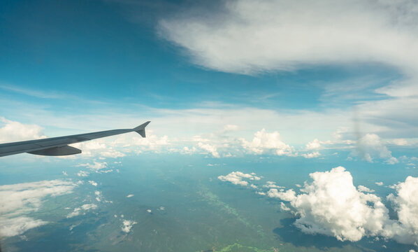 View from inside plane through plane window over blue sky, white clouds and green mountain. Commercial airline flight for summer travel. Plane wing above the city. Airplane flying above land.
