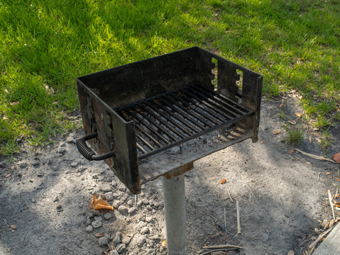 A heavy-metal barbecue grill, containing burned charcoal ash, stands securely mounted on a steel pole in an outdoor public recreation area.