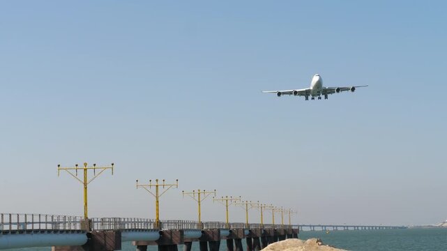 Four-engine passenger airplane approaching for landing at airport over sea water with runway lights. Clear blue sky, bright daylight, wide angle shot of aircraft arrival.