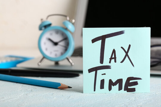 Laptop with alarm clock, stationery and text TAX TIME on grunge blue table near white wall