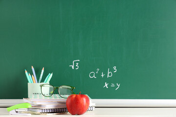 Obraz premium Apple, eyeglasses and stationery on desk near chalkboard with formulas. Back to school concept
