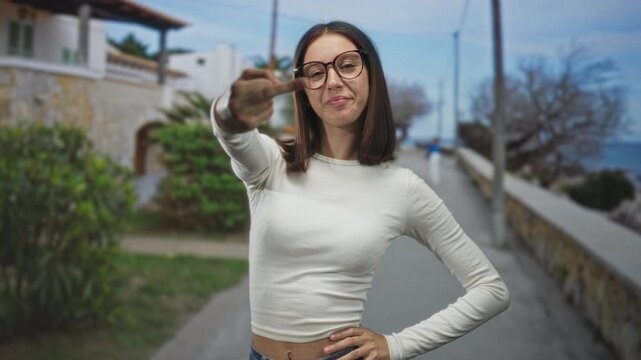 Young woman in white shirt flips middle finger to camera on a seaside street promenade, hand extended and smirking; rebellion defiance.