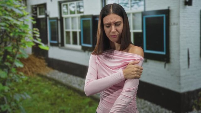 Woman in a pink long sleeve shirt rubbing her upper arm with a pained grimace standing outside in front of a white building with shuttered windows; discomfort introspection.