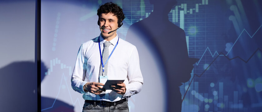 Smiling young male speaker with headset and tablet presenting financial data on a screen