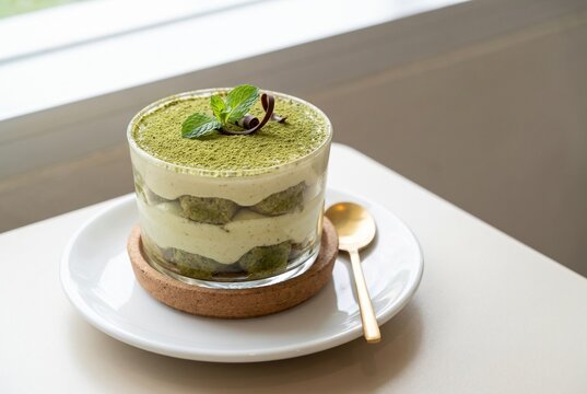 ガラスカップに重ねられた抹茶クリームとスポンジの層。抹茶パウダーとミントの飾り、木のコースターと自然光が、抹茶スイーツの魅力を丁寧に伝える一枚