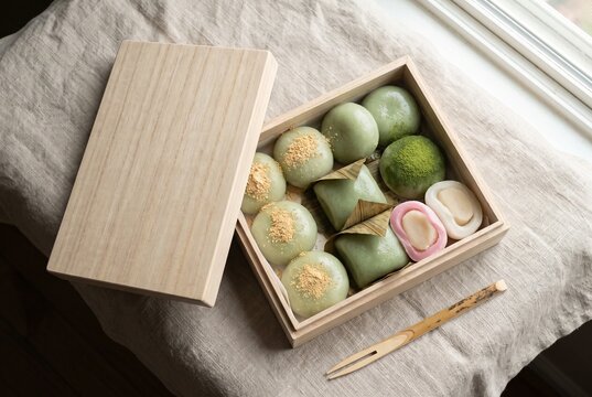 木箱に丁寧に並べられた和菓子。抹茶の菓子や葉包みの餅、ねじり菓子が自然光に照らされ、和の繊細な美しさと職人の手仕事を感じさせる一枚