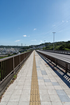 快晴の空と左側と正面の斜面に住宅地のある橋の点字ブロックが続く歩道と車道