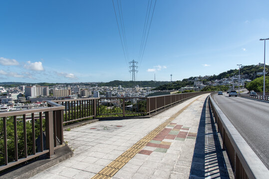 快晴の空と左側と正面の斜面に住宅地のある橋の点字ブロックが続く歩道と車道