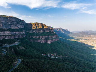 Fototapeta premium A panoramic landscape featuring layered rocky cliffs, dense green valleys, and a winding road under a clear blue sky. Small settlements nestle in the forested slopes.