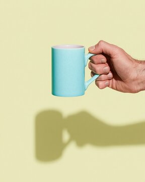 Blue ceramic mug mockup held by a single hand against a pale yellow background, showcasing a smooth satin glaze and sharp shadows for beverage branding and advertising