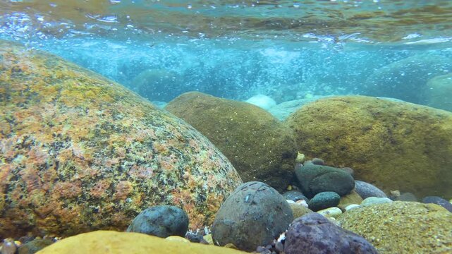 Colorful river stones sit on the streambed as clear water and bubbles flow past in a shallow underwater shot.