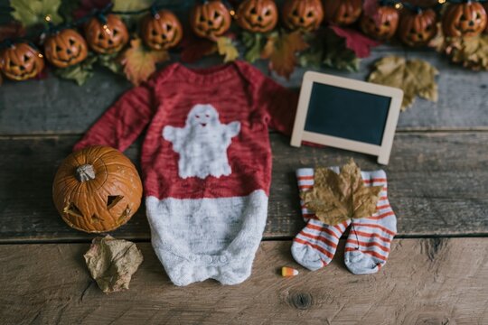 Red ghost print baby baby bodysuit and pumpkin flat lay on a rustic wooden surface for a cozy Halloween pregnancy announcement and seasonal greeting