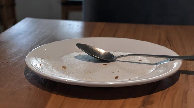 An empty white ceramic plate with food crumbs, spoon, and fork after finishing a meal on a wooden restaurant table, shot in slow motion.
