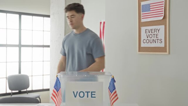 Man places ballot into clear ballot box beside american flags on table in polling building; civic pride.