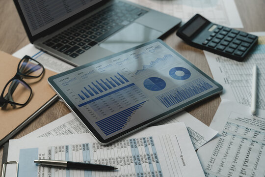 business project finance investment dashboard beside laptop, calculator, eyeglasses, pen, printed reports supporting financial analysis, document reporting, business finance workflow.