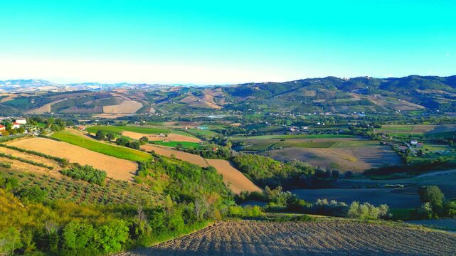 Gorgeous aerial pedestal up push-in shot from Monte Rinaldo, sweet rolling Marche hillscapes in checkerboard pattern interspersed with lawns, thickets, brown fields, buildings, under a gradient sky