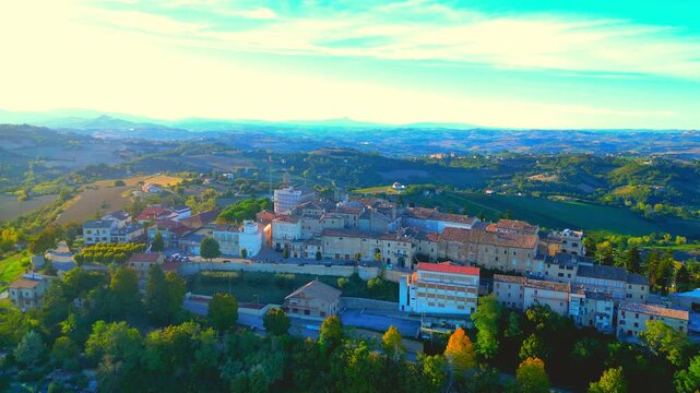 Careful aerial pedestal down shot from Monte Rinaldo, descending views of the historic town centre on a vegetation-filled promontory amongst Marche hillscapes, creamy clouds, azure sky, blue mountains