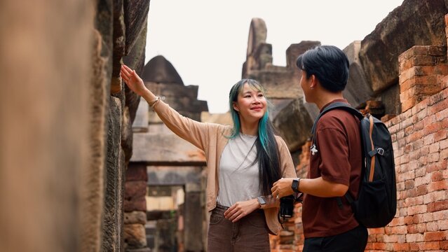 Young Asian couple walking and exploring together at ancient stone temple ruins. Happy tourists enjoying cultural heritage discovery, summer vacation holiday and travel lifestyle adventure.