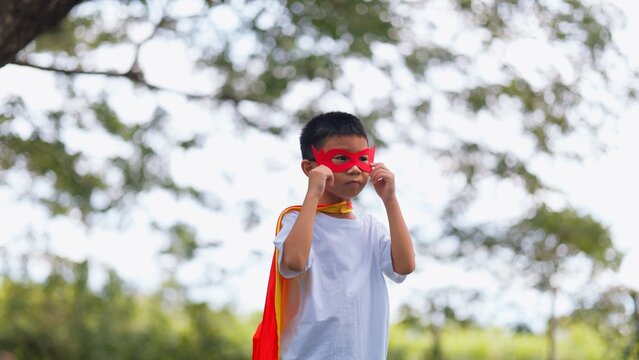 Happy Asian children playing superhero roleplay with colorful cape and mask running in green park. Kids imagination, creative future dream, and brave hero inspiration concept.
