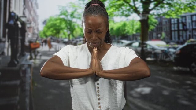 Woman with hands pressed together in prayer gesture and closed eyes, tense face, visible palms and forearms on an urban street lined with trees and parked cars; grief seeking solace.