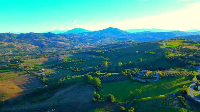 Pristine aerial truck left shot from Monte Rinaldo, balmy afternoon sunlight shining on winding roads perched on green hills, interspersed with brown fields, thickets, far rugged Sibillini mountains