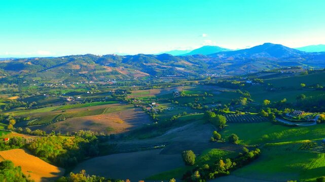 Cinematic aerial push-in pan right shot from Monte Rinaldo, from Marche valleys with thickets and fields, to the nearby hills, reaching distant Sibillini mountains in all their majestic, hazy splendor