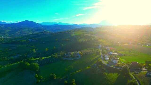 Delightful aerial push-in shot from Monte Rinaldo, the nearby countryside imbued with the precious light of the afternoon sun, having roads, cars, buildings, fields, meadows, blue Sibillini mountains