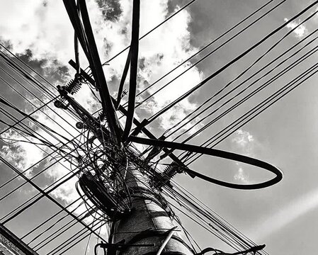 Black and white power lines and electric pole with tangled utility cables against a cloudy sky. Dramatic urban infrastructure and electricity distribution network.
