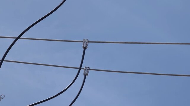 Pan down video of T-clamps connecting jumper wires to high voltage bare aluminum cables, industrial electrical power grid distribution network infrastructure against a blue sky.