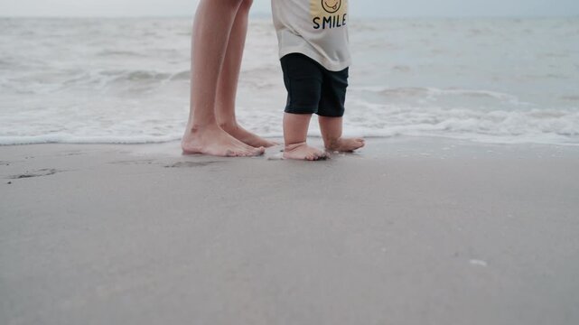 Mother and baby walking barefoot on beach sand during summer family vacation.