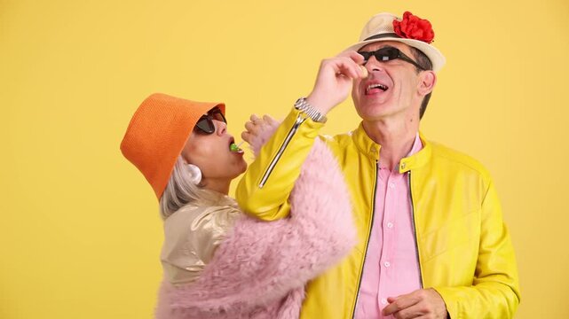 Playful senior couple flirting with lollipops on yellow background