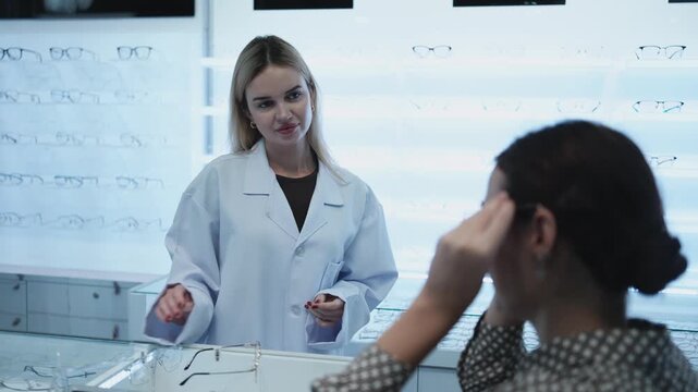 White female optometrist helps and guides customers to choose eyeglasses for improved eyesight with individual lifestyles, consults on decisions, and options choices at store display in optical shop.