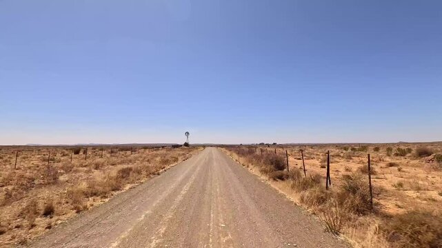 Vehicle driving along long dirt road in dry landscape with metal windmill in South African desert.