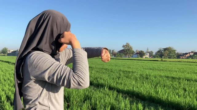 Focused young Muslim woman in a hijab doing shadow boxing exercises outdoors in a vibrant green rice field on a clear sunny morning.