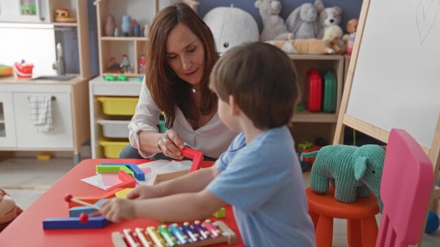 Woman teacher guides boy holding mallet over wooden xylophone on red table in preschool classroom; nurturing learning.