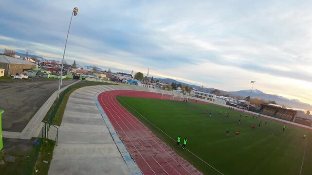 Players training at Estadio Municipal Victor Borquez Miranda surrounded by an athletics track and empty bleachers with mountains and sunset in the distance at Puerto Natales Chile, FPV aerial